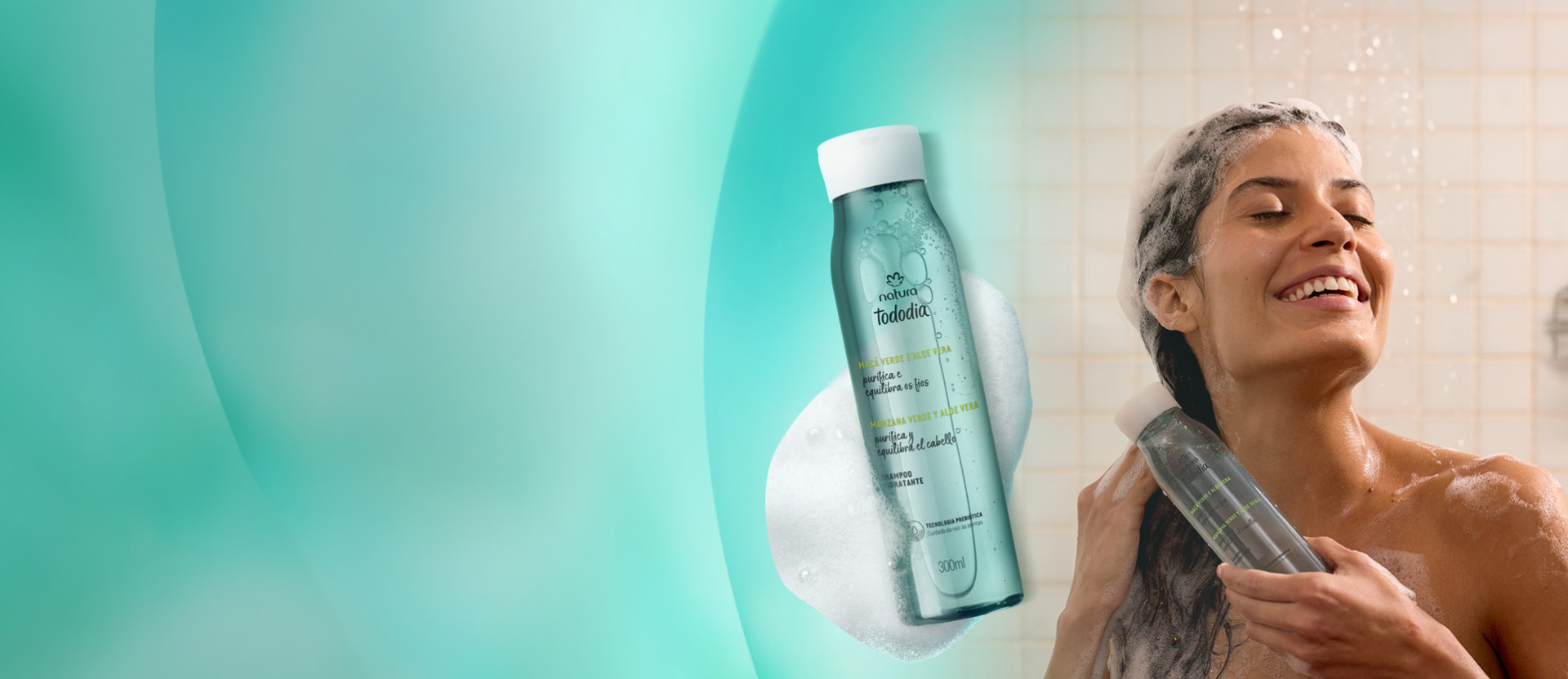 Mulher sorrindo e lavando o cabelo no chuveiro, com o rosto molhado e espuma no cabelo. À frente dela, um shampoo Natura Tododia Maçã Verde e Aloe Vera na cor verde-água, com espuma ao redor.
