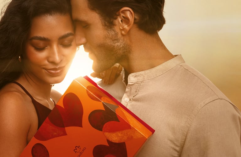 Foto de um casal se beijando, com uma caixa de presente vermelha com corações nas mãos, em fundo desfocado em tons quentes.