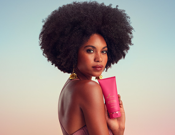 Modelo negra de cabelos crespos usa vestido longo rosê e brincos dourados. Ela olha para a câmera e exibe o Desodorante Hidratante Iluminador Corporal Perfumado Luna Divina.