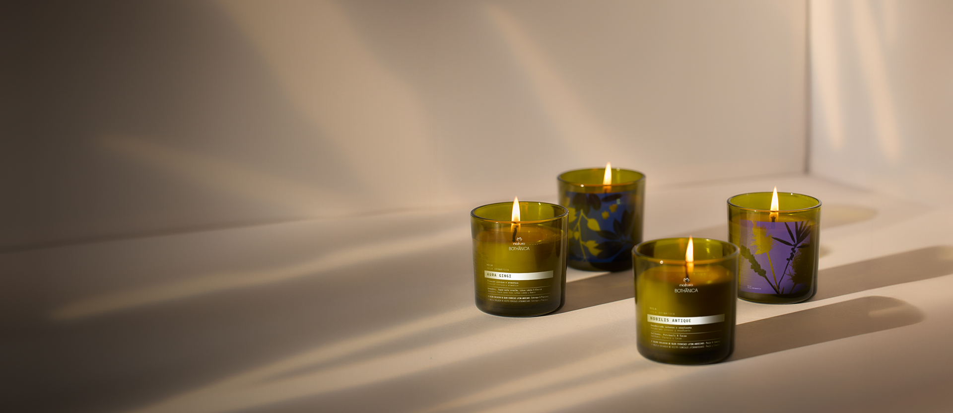 Fundo em tons claros com fachos de luz, quatro velas acesas da linha Natura Bothânica à direita.