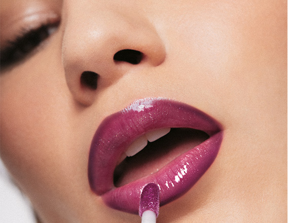 Mulher de pele branca aplica um gloss roxo brilhante sobre os lábios delineados em tom magenta. Nos olhos, ela usa maquiagem esfumada em tons de marrom.