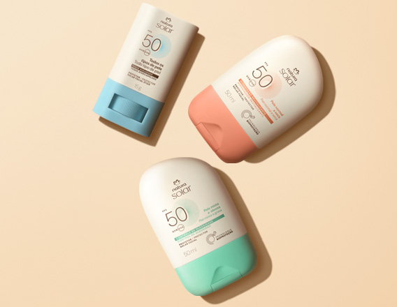 Três Protetores Solares Faciais FPS 50 Natura dispostos em diagonal. Todos têm embalagem branca, mas a base do stick é azul e as tampas dos demais são verde e laranja. Fundo em tom de creme.