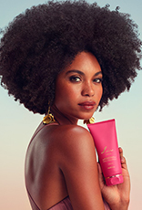 Modelo negra de cabelos crespos usa vestido longo rosê e brincos dourados. Ela olha para a câmera e exibe o Desodorante Hidratante Iluminador Corporal Perfumado Luna Divina.
