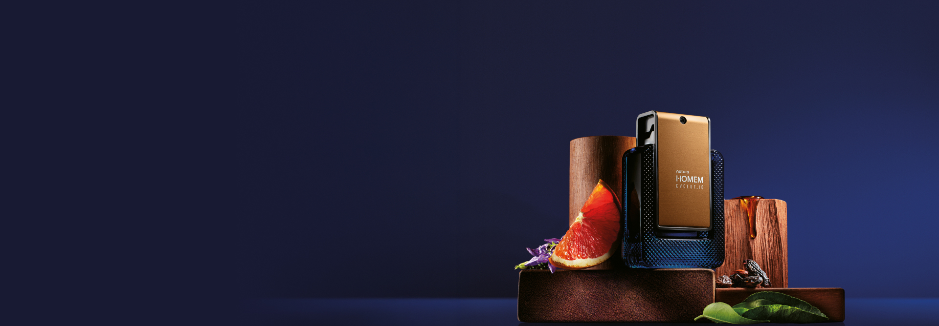 Frasco da fragrância Natura Homem Evolut.io sobre madeiras e cercado de frutas cítricas, flores, folhas e sementes. Fundo azul-escuro.