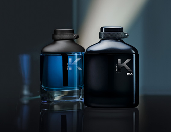 Frascos das fragrâncias K em azul e K Max em preto. Fundo escuro com reflexos e um feixe de luz suave ao fundo.