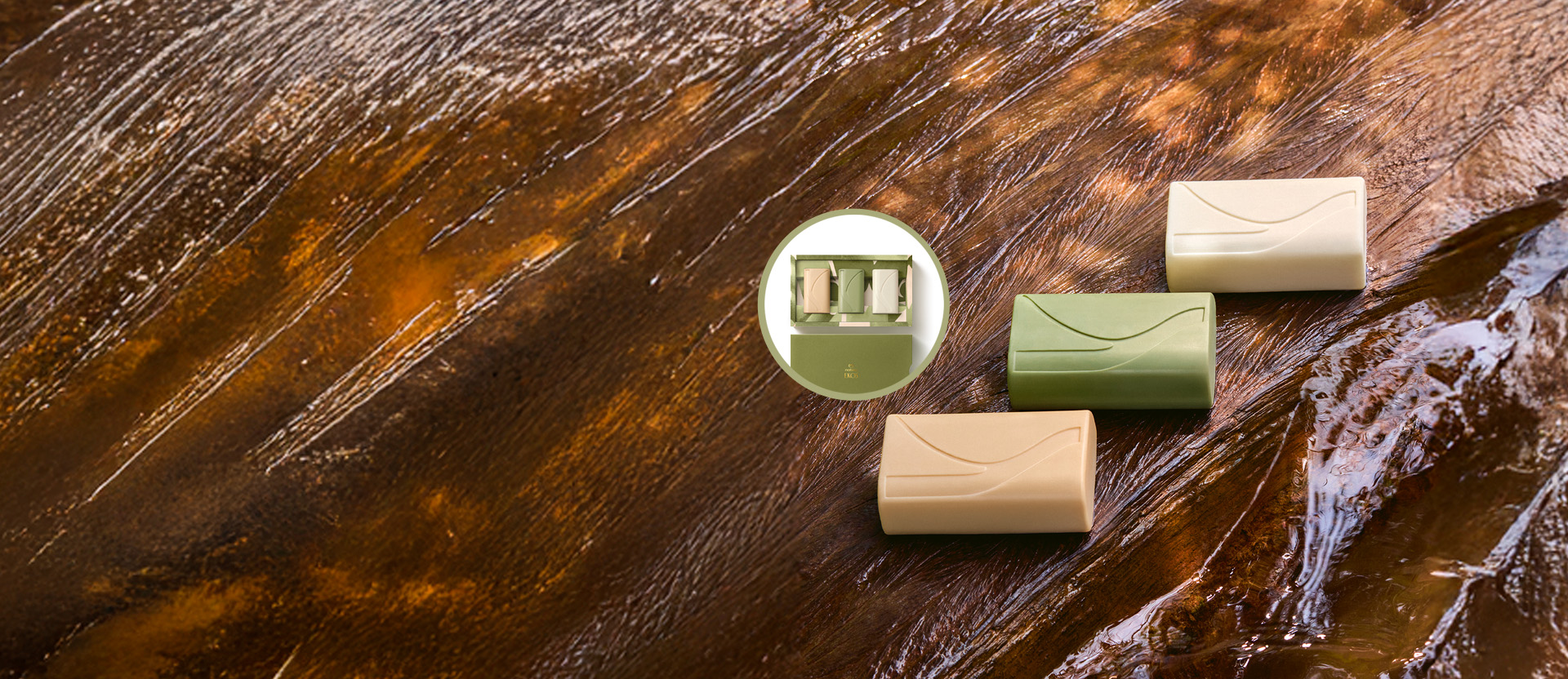 Três Sabonetes Natura Ekos Ryos, em tons bege, verde e branco, estão sobre uma superfície de madeira molhada. No centro, há um detalhe em círculo mostrando a caixa de presente com os três sabonetes.