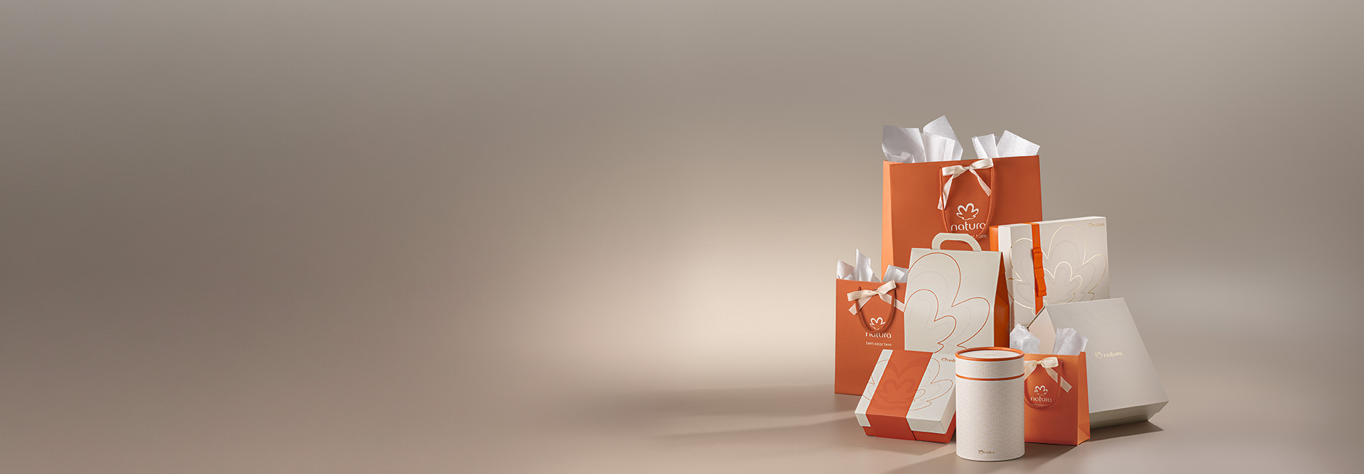 Conjunto de sacolas e caixas de presente da Natura em tons de laranja, bege e branco, organizadas sobre um fundo neutro.