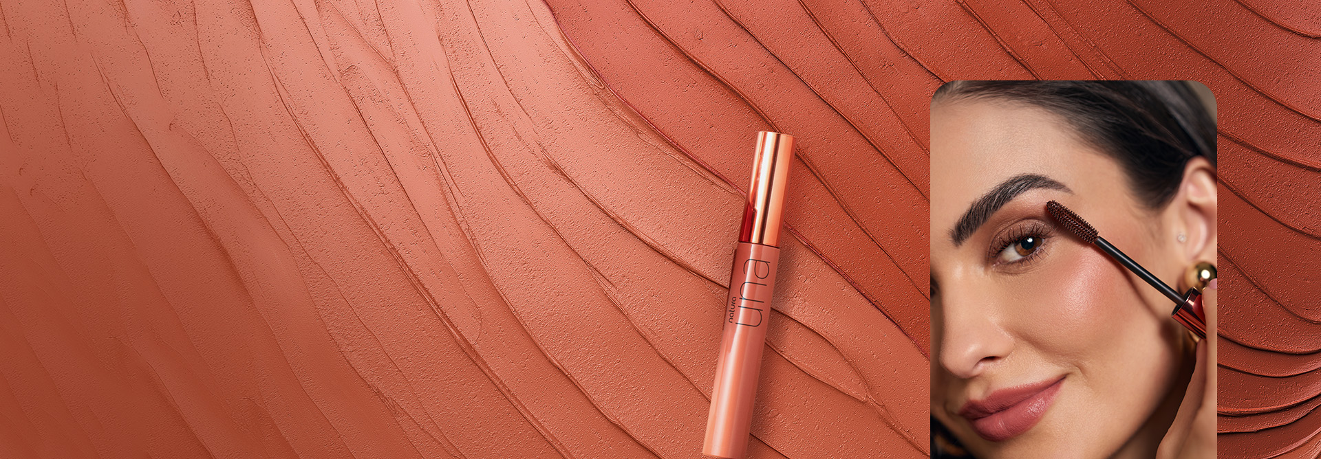 Tubo nude metalizado da Máscara para Cílios Volume Marrom Una, em um fundo com produto cremoso rosado espalhado em ondas. Ao lado, mulher branca de cabelo escuro aplica a máscara nos cílios.