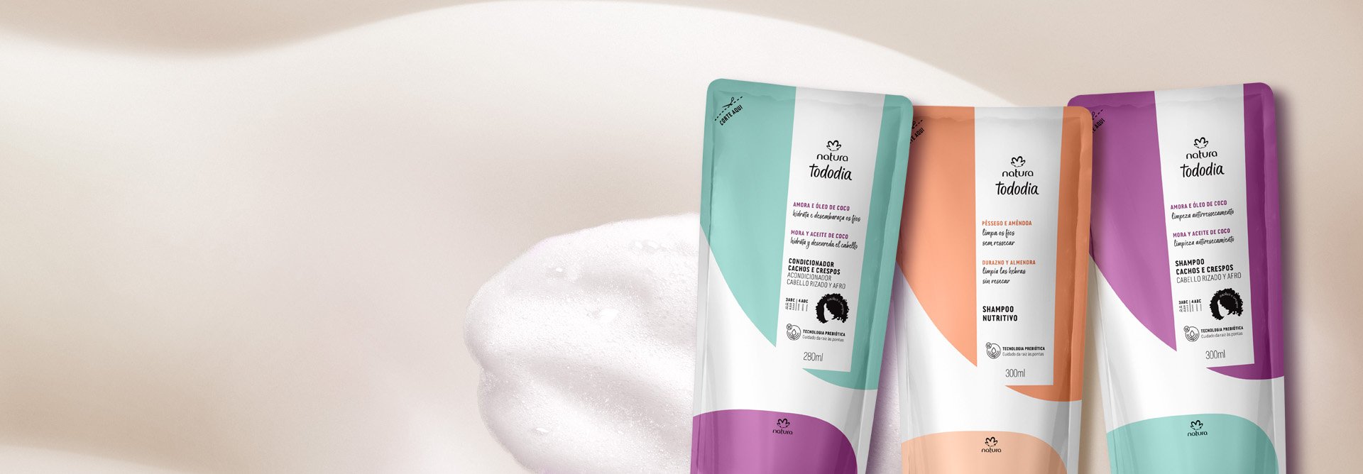 Sachê laranja do Shampoo Nutritivo Tododia Pêssego e Amêndoa e 2 sachês roxos com azul, um do Shampoo e outro do Condicionador Tododia Amora e Óleo de Coco. Estão em fundo bege com espuma branca.