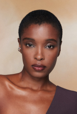 Close de rosto de mulher com pele negra, cabelo curto e maquiagem discreta; ombro nu visível. Fundo bege suave, olhar direto à câmera.
