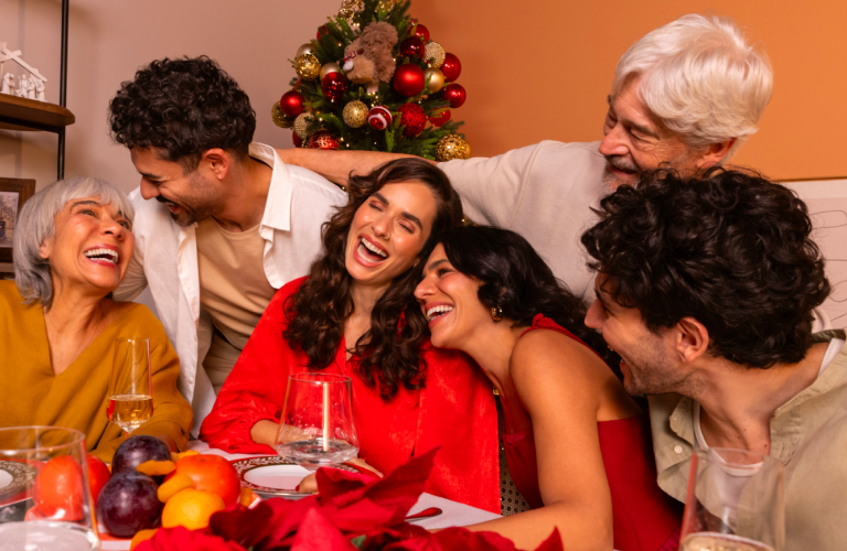 Foto em ambiente interno de sete pessoas reunidas e sorrindo ao redor de uma mesa decorada, com uma árvore de Natal ao fundo.