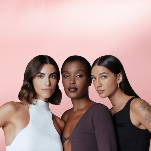 Três mulheres: uma com top branco, cabelo castanho curto e pele branca, uma com blusa marrom, pele negra e cabelo curto, e uma com top preto, cabelo longo liso e pele branca. Fundo degradê rosa-claro.