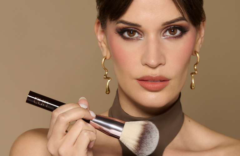 Mulher com maquiagem elaborada segurando um pincel de blush e um batom, com brincos dourados e lenço marrom ao pescoço. Fundo bege.