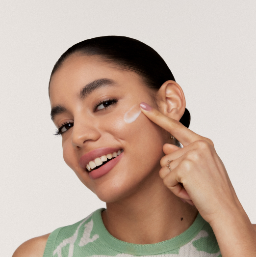Modelo morena de cabelos presos e blusa verde e branca sorri para a câmera enquanto aplica um produto da linha Natura Faces Controle de Oleosidade na maçã do rosto com o dedo indicador.