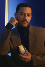 Homem de cabelos curtos, usando camisa de gola alta preta e terno marrom, segura a fragrância Natura Homem Evolut.io próximo ao corpo, em fundo azul e moldura branca iluminada.