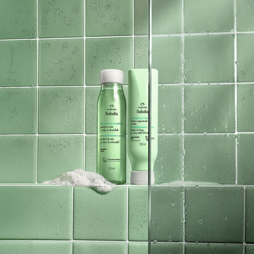 Frascos verdes com tampas brancas de Shampoo e Condicionador da linha Tododia Chá de Hortelã e Gengibre. Eles estão sobre uma espuma branca, em um banheiro com azulejos verdes e box de vidro.