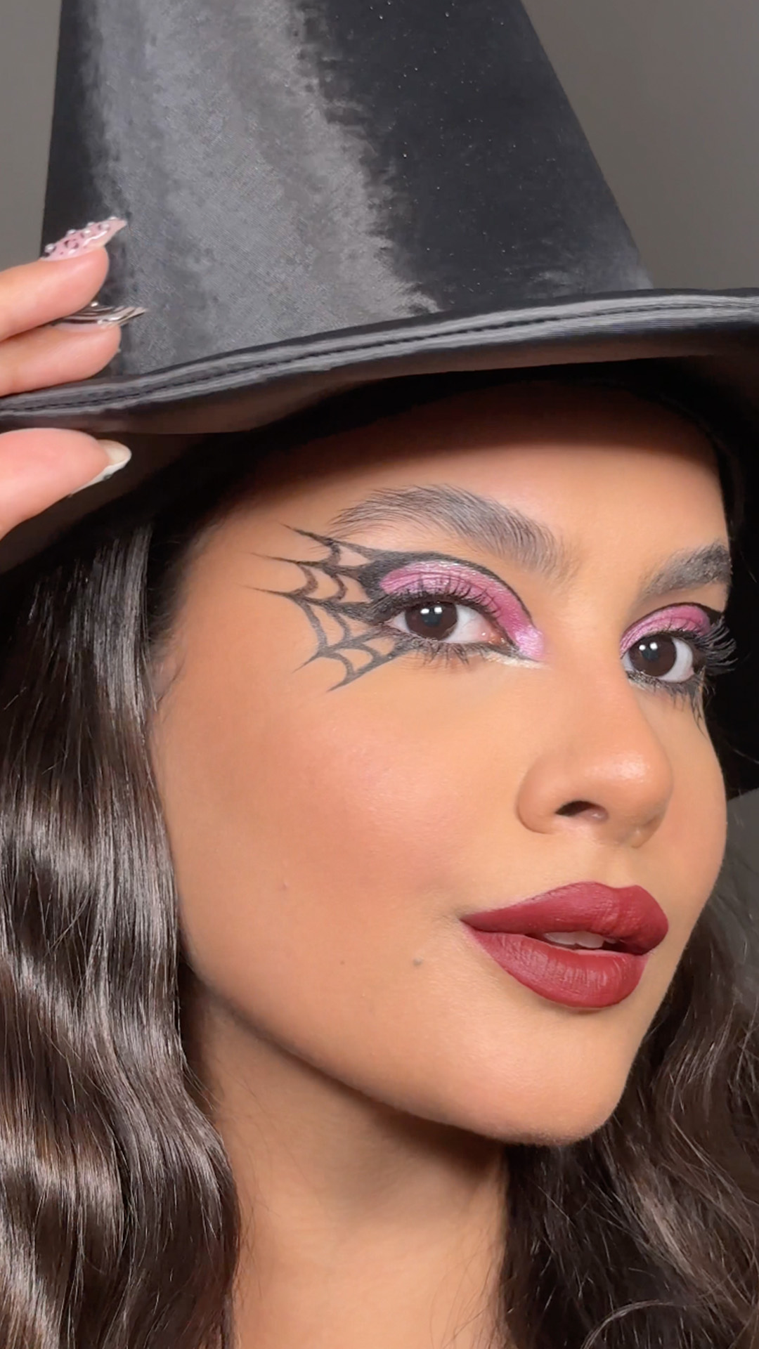 Um tutorial de maquiagem de Halloween estilo "Bruxa". Uma jovem usa lápis preto para delinear o côncavo e aplica sombra roxa retrátil ("púrpura mistery") na pálpebra. Ela usa delineador preto para fazer um gatinho e uma teia de aranha detalhada no canto externo do olho. O visual é finalizado com batom líquido mate marsala ("marsala night"). Na tela, aparece a jovem usando um chapéu de bruxa e o texto: "make de bruxa".