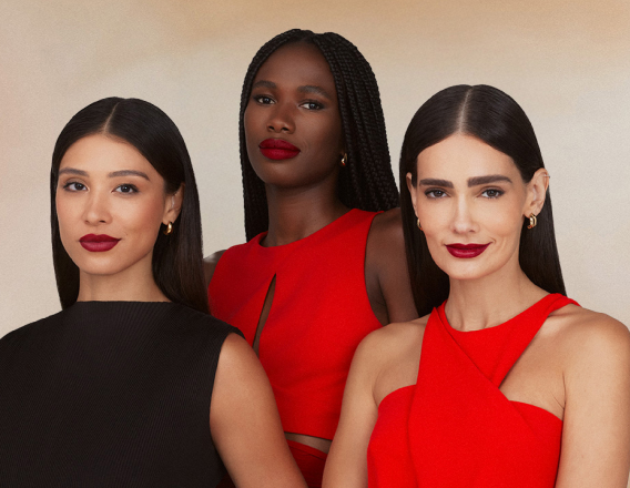 Três mulheres com diversos tons de pele usando batom vermelho intenso; duas vestem roupa vermelha e uma preta. Fundo neutro bege-claro, expressão serena e joias discretas.