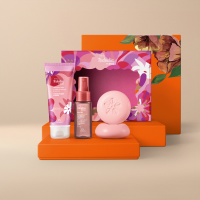 Presente Tododia Flores Essenciais, incluindo 1 Body Splash, 1 Creme Desodorante Nutritivo para o Corpo, 2 Sabonetes em Barra e 1 Caixa Presenteável. Embalagens em tons de rosa em fundo bege-claro.