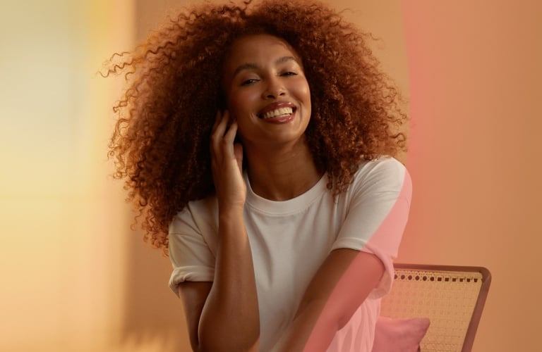 Mulher com as pernas cruzadas e o cotovelo direito apoiado nas pernas, enquanto passa a mão no pescoço e sorri