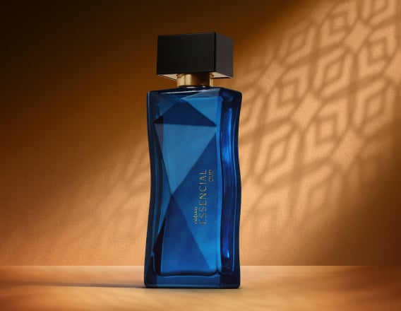 Deo Parfum Essencial Oud Feminino em frasco facetado azul com tampa preta. Está em fundo cor areia com sombra geométrica projetada.