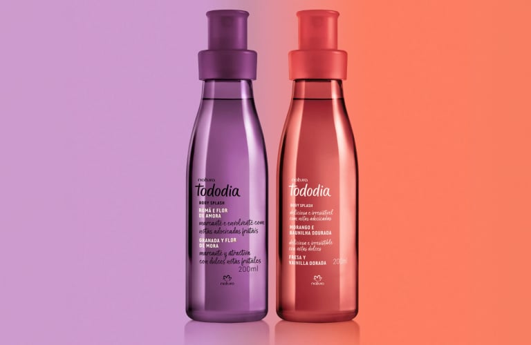 Foto com fundo degradê lilás e vermelho. Frascos de Body Splash Tododia Romã e Flor de Amora e Morango e Baunilha Dourada lado a lado.
