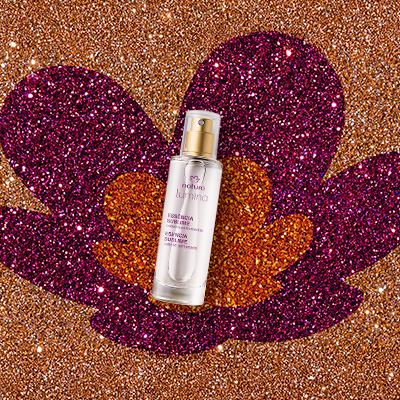 Frasco transparente com tampa dourada da Essência Sublime Lumina. Produto em destaque sobre fundo em glitter laranja com a flor símbolo da Natura aplicada em glitter laranja e rosa.