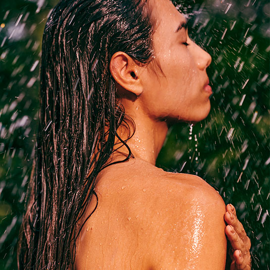 Pessoa de pele morena e cabelos longos e escuros está de olhos fechados sob a água, com gotas escorrendo pelo corpo. O fundo desfocado é verde, sugerindo natureza ao redor.