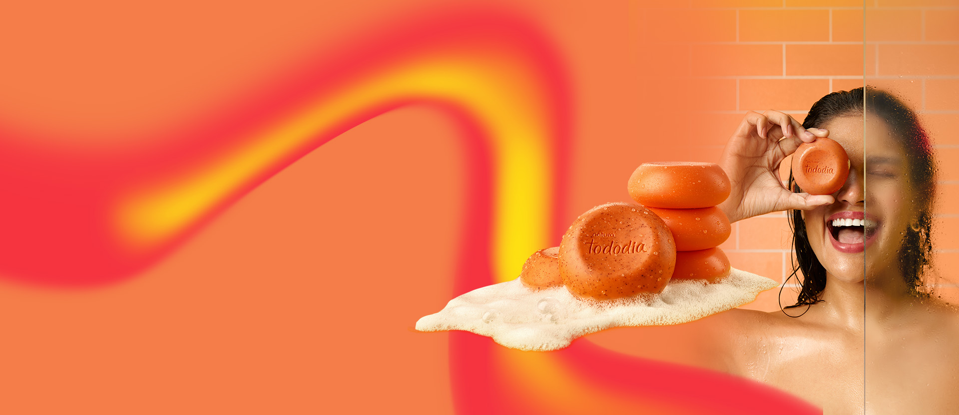Sabonete em Barra Esfoliante Tododia Energia em destaque sobre fundo laranja e ondas avermelhadas. Junto a ele, mulher sorri de olhos fechados e exibe o sabonete à frente do olho direito.