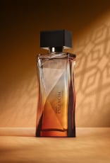 Deo Parfum Essencial Mirra Feminino em frasco facetado degradê âmbar com tampa preta. Está em fundo cor areia com sombra geométrica projetada.