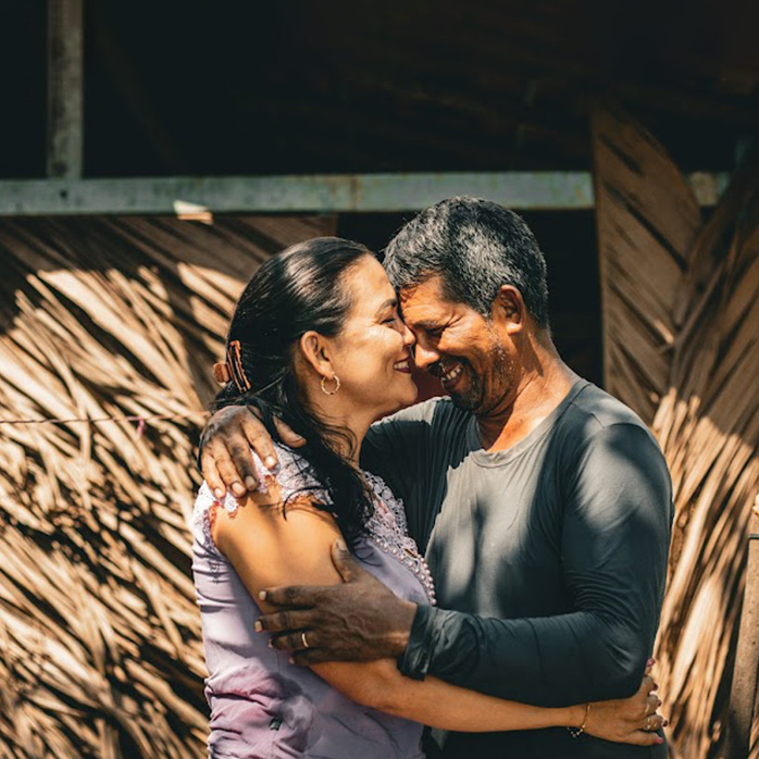Casal ribeirinho abraçado à frente de casa com palhas naturais.