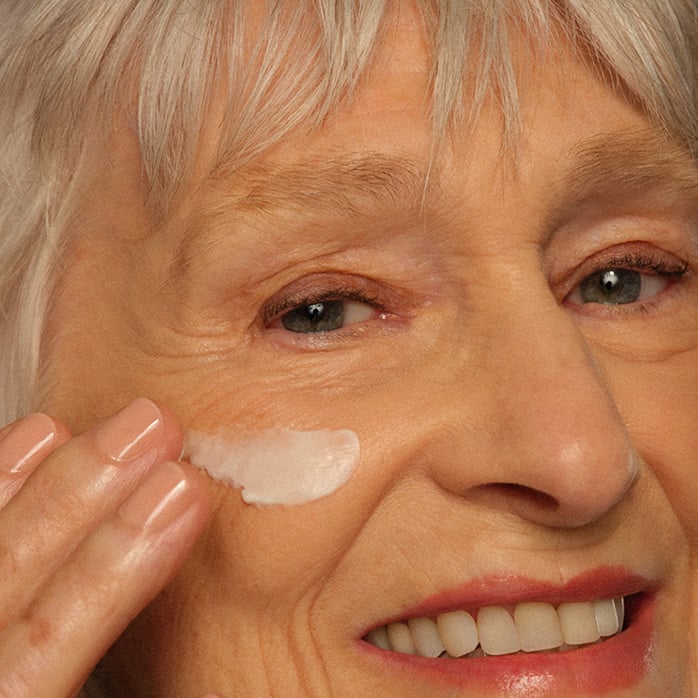 Close do rosto de uma mulher idosa sorrindo, com cabelos grisalhos. Ela aplica uma pequena quantidade de creme facial branco na bochecha com a ponta dos dedos, destacando as linhas de expressão naturais e a textura da pele.
