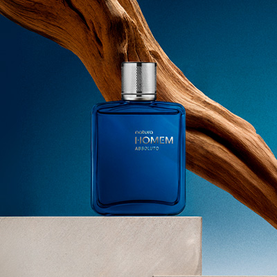 Deo Parfum Natura Homem Absoluto, com frasco de vidro transparente azul-médio e tampa prateada, destaca-se sobre superfície clara e fundo azul com madeiras nobres que compõem a fragrância.