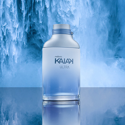 Desodorante Colônia Kaiak Ultra Masculino com frasco de vidro em gradiente azul e tampa prateada. O produto está sobre superfície reflexiva azul com uma cachoeira ao fundo.