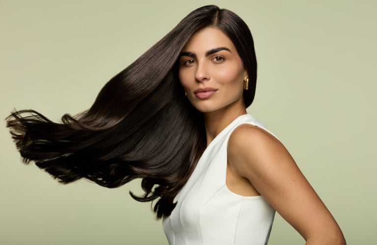 Foto de uma mulher com cabelo liso escuro ao vento. Ao fundo, cor verde claro. Ela veste blusa branca sem mangas e usa brincos dourados.