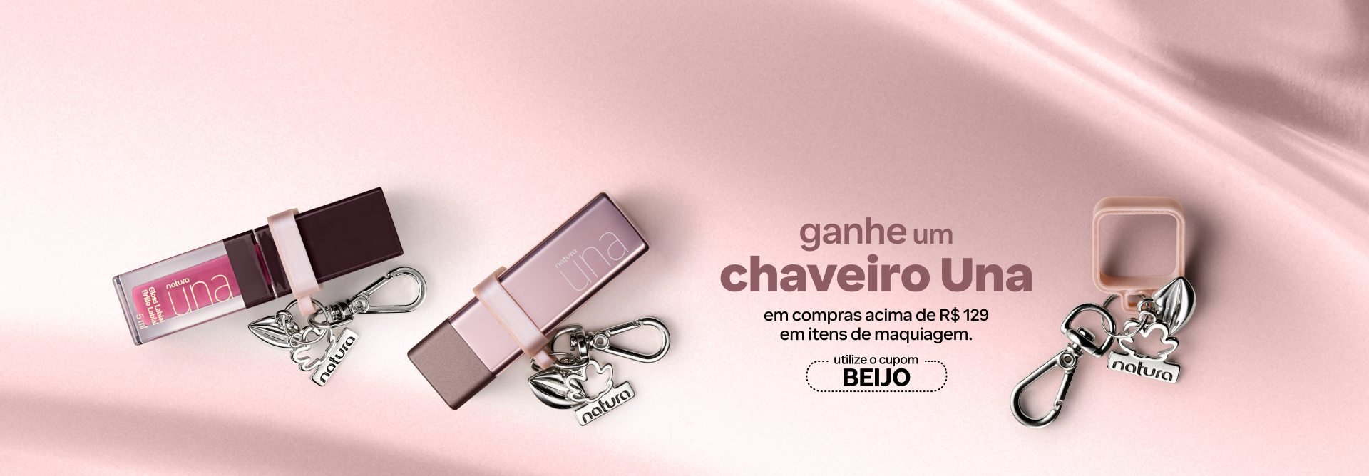Batons Natura Una com chaveiros prateados e o texto: ganhe um chaveiro Una em compras acima de R$ 129 em itens de maquiagem. Utilize o cupom BEIJO. Fundo com textura em tons rosados.