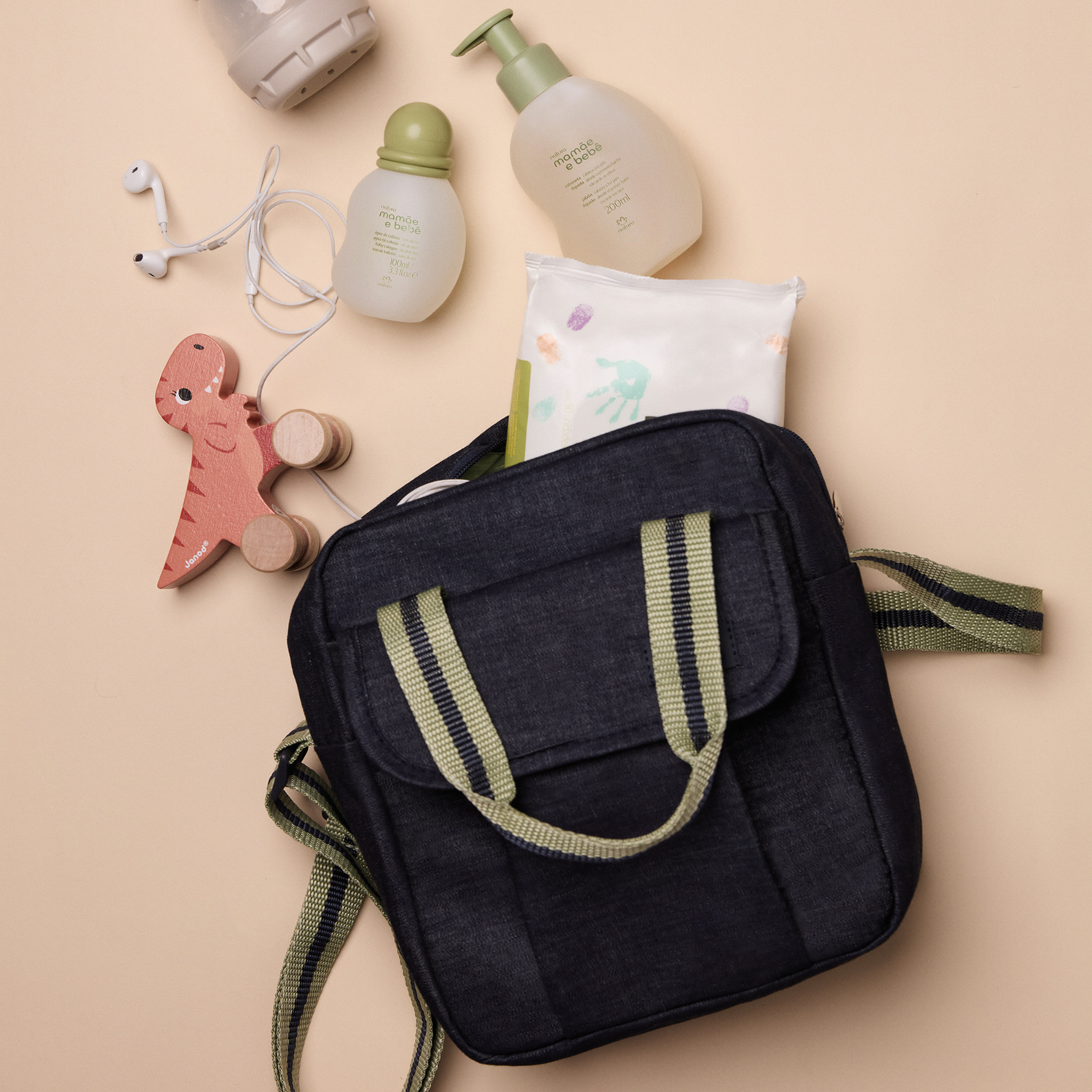 Bolsa jeans Mamãe e Bebê aberta sobre superfície bege. Dentro há um pacote de lenços umedecidos. Outros objetos estão ao redor, como colônia, fones de ouvido, mamadeira e um dinossauro de brinquedo.