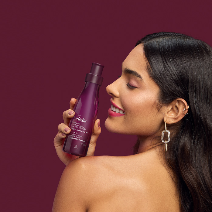 Mulher de cabelos castanhos segura produto Natura Tododia em fundo vinho.