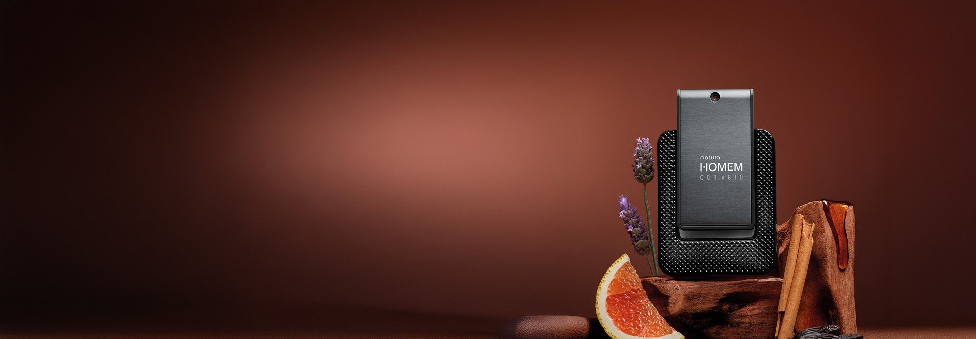 Deo Parfum Natura Homem Cor.agio em frasco cinza-escuro com base preta texturizada. Está sobre madeira escura, cercado por flores roxas, canela e uma fatia de laranja, em fundo marrom.