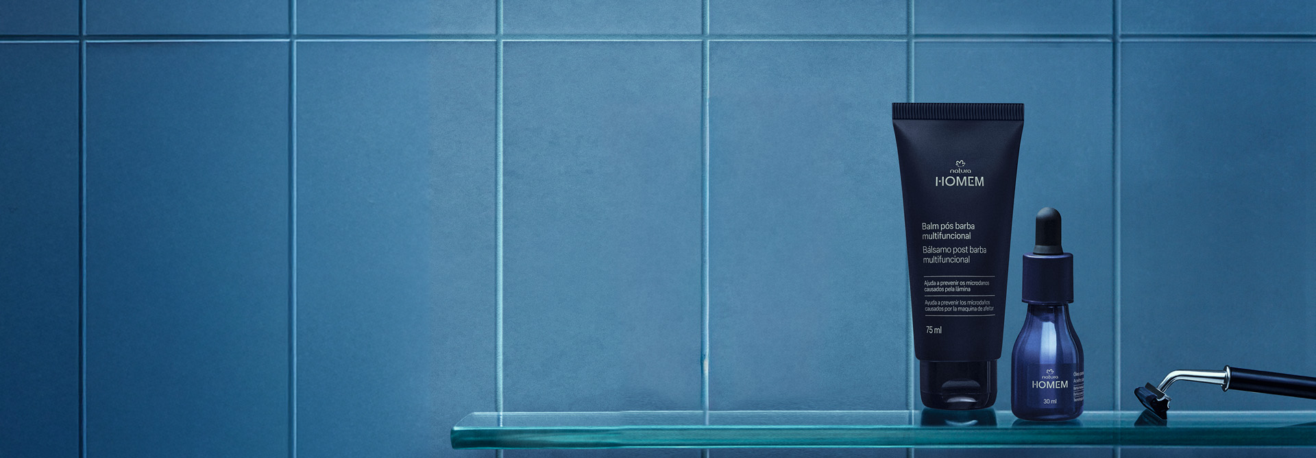 Natura Homem Balm Pós-Barba Multifuncional e Óleo para Barba, com frascos azul-escuros e tampas pretas, destacam-se em prateleira de vidro em fundo azulejado azul-celeste. Ao lado, lâmina de barbear.