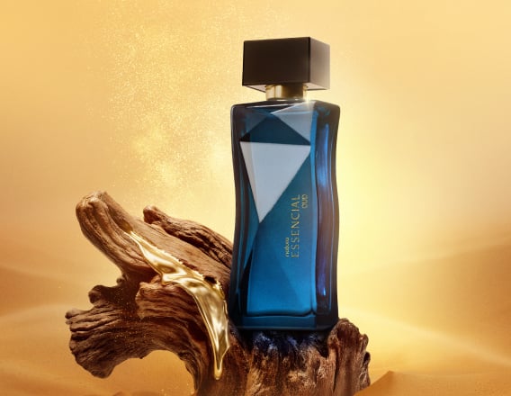 Frasco de Essencial Oud Feminino, em tom de azul-escuro e tampa preta, aparece em destaque no centro da tela sobre fundo multifacetado dourado, fazendo alusão ao design do frasco de Essencial.