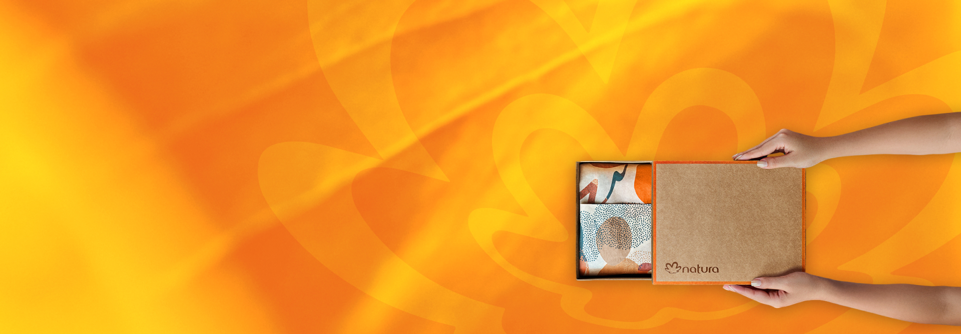 Na imagem, mãos femininas abrem um presente Natura, com caixa parda, logo Natura, detalhes em laranja e papel de presente decorado. Fundo em tons de amarelo e laranja com a flor-símbolo da Natura.