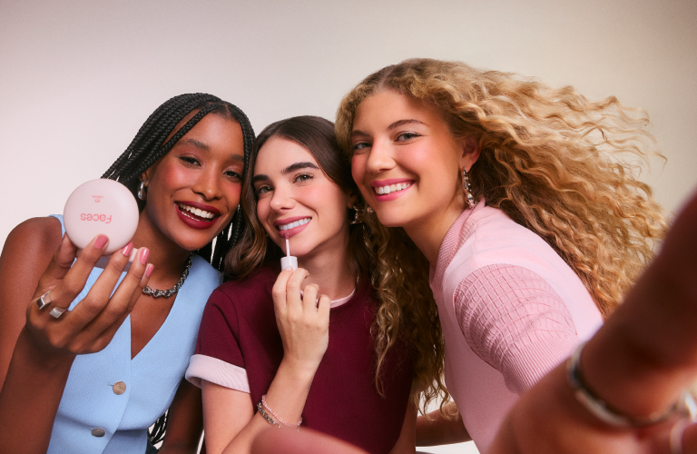 Três amigas sorrindo juntas em uma selfie, uma delas segurando um pote de pó compacto Faces e outra aplicando um gloss labial.