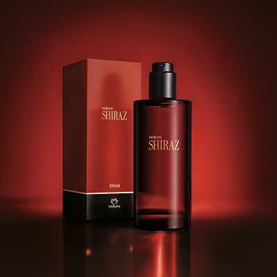 Desodorante Colônia Shiraz com frasco de vidro vermelho-escuro ao lado da caixa vermelha com detalhes em preto. Ambos estão sobre superfície reflexiva escura, com fundo em degradê de vermelho.