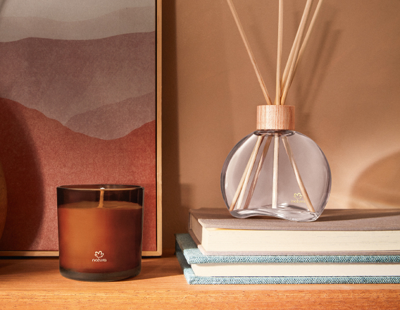 Vela Perfumada e Difusor Natura 861 Guaiaco Pataqueira aparecem em destaque sobre prateleira de madeira. O Difusor repousa sobre dois livros e a Vela Perfumada destaca-se à frente de tela abstrata.