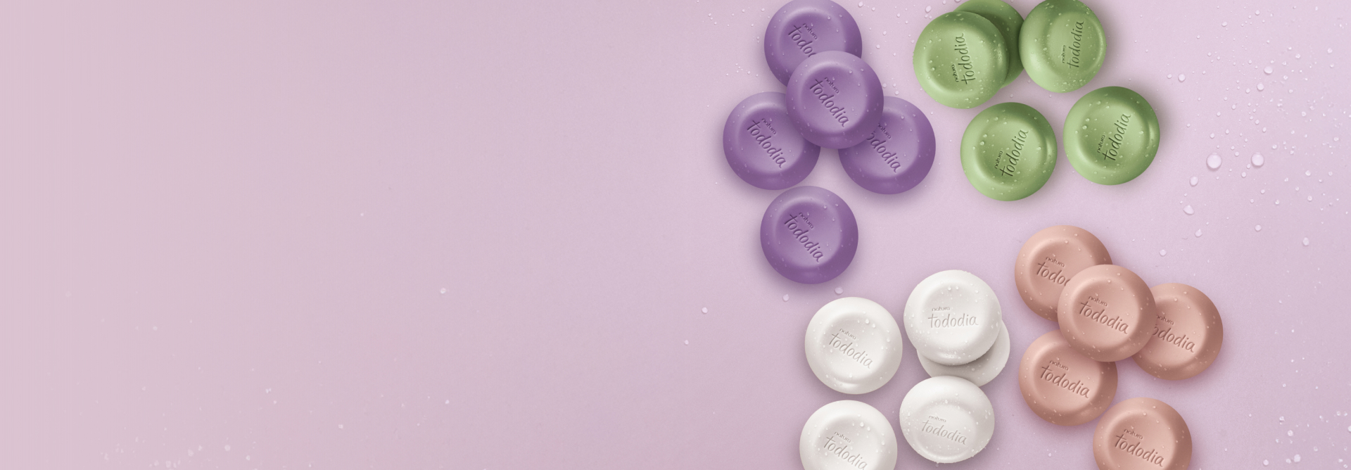 Conjunto de sabonetes redondos da linha Tododia em fundo roxo com gotas de água na superfície. Sabontetes nas cores branco, verde, rosa e roxo.