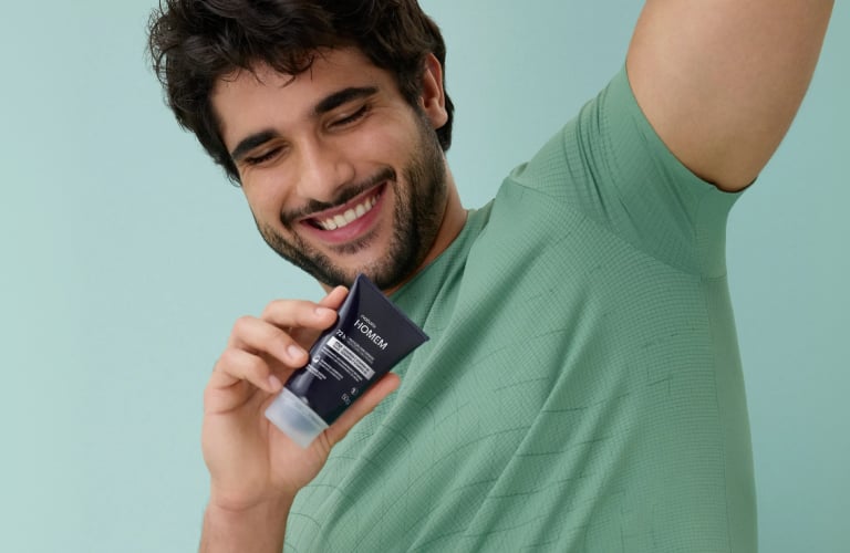 Foto de homem sorrindo com camiseta verde, fundo claro e produto Natura Homem na mão.
