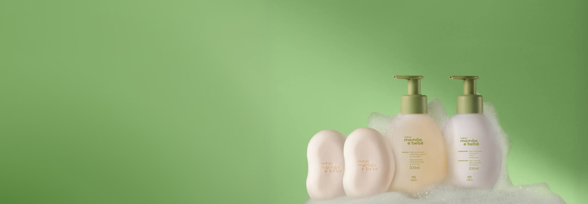 Kit Mamãe e Bebê em fundo verde-claro, contendo 2 sabonetes em barra de cor bege, 1 shampoo e 1 condicionador. Frascos brancos com tampa pump verde. Os produtos estão envolvidos por uma espuma branca.