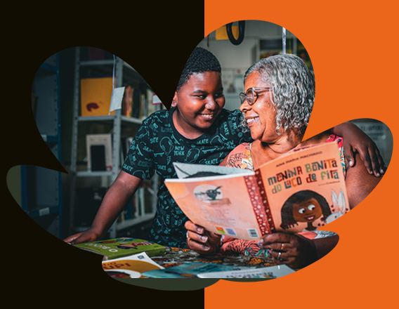 Imagem de uma senhora sorridente de cabelos grisalhos e óculos, lendo um livro com o título "Menina Bonita do Laço de Fita" em voz alta para um jovem com um sorriso, que a abraça gentilmente por trás. Ambos estão em uma biblioteca, com estantes de livros ao fundo, e a cena é emoldurada por um fundo em preto e laranja.