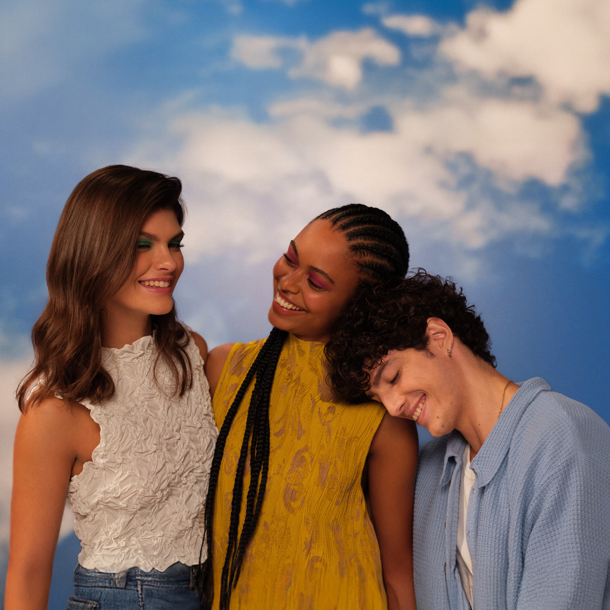 Três jovens, duas mulheres e um homem, aparecem abraçados e sorrindo sob um fundo de céu com nuvens. Estão maquiados com cores vibrantes e expressam alegria.